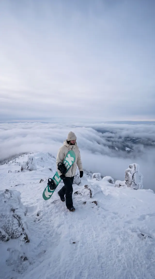 A snowboarder carrying a teal board along a snowy ridge