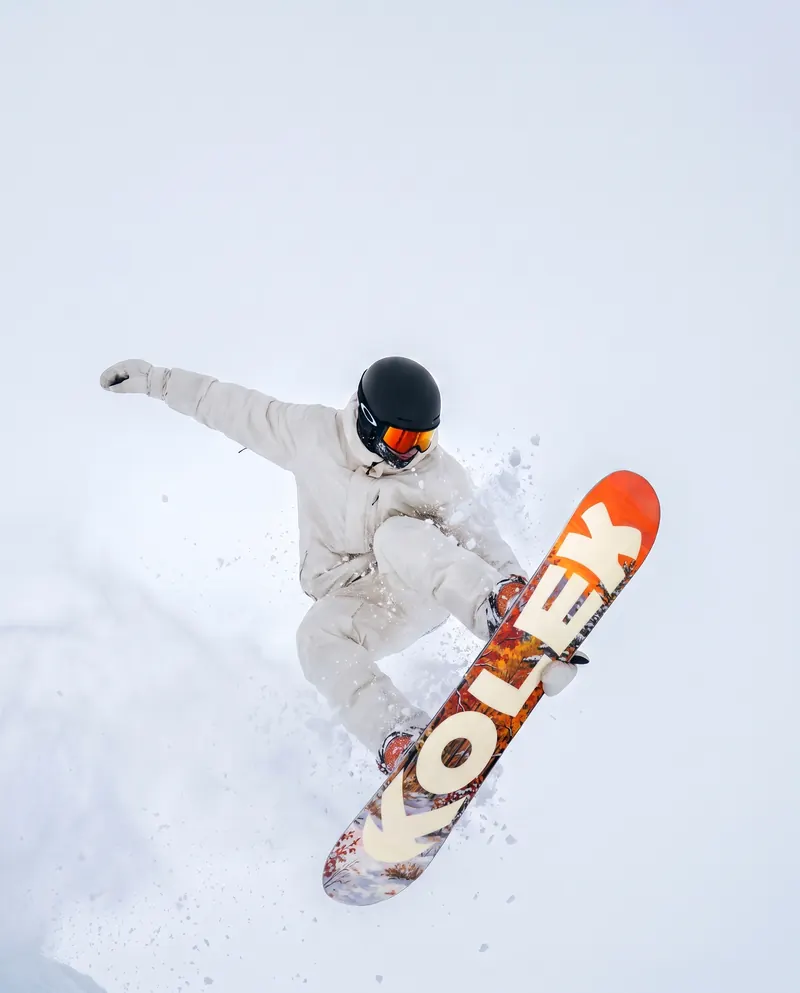 A snowboarder in a cream jacket mid-air, grabbing an orange Kolek board